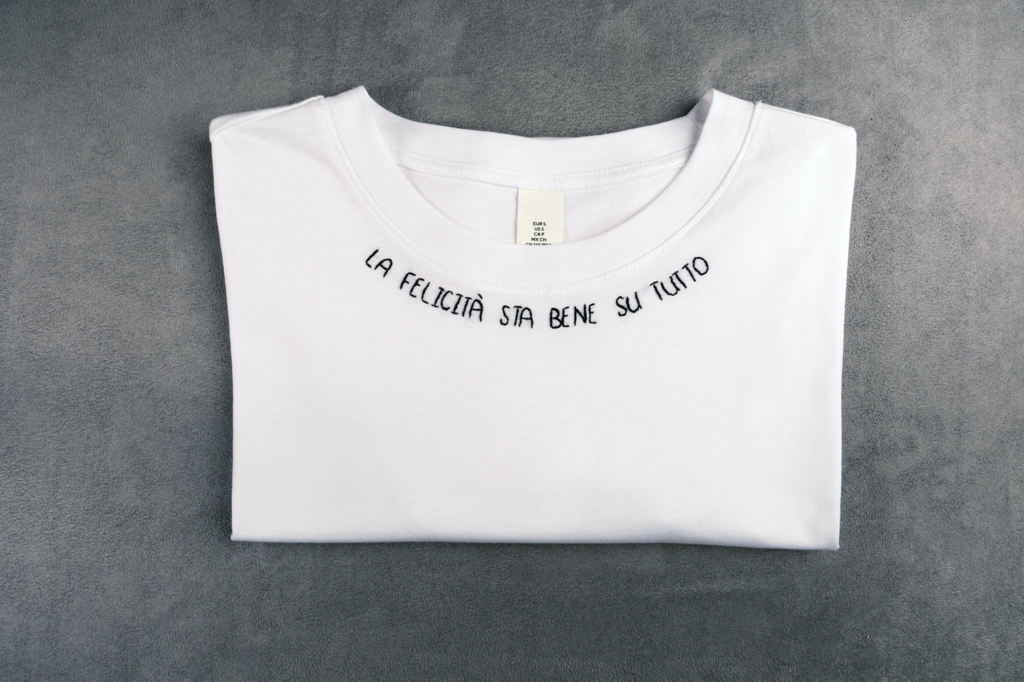 Dettaglio T-shirt “LA FELICITÀ STA BENE SU TUTTO”, la maglietta in 100% cotone è ricamata con la scritta "la felicità sta bene su tutto"