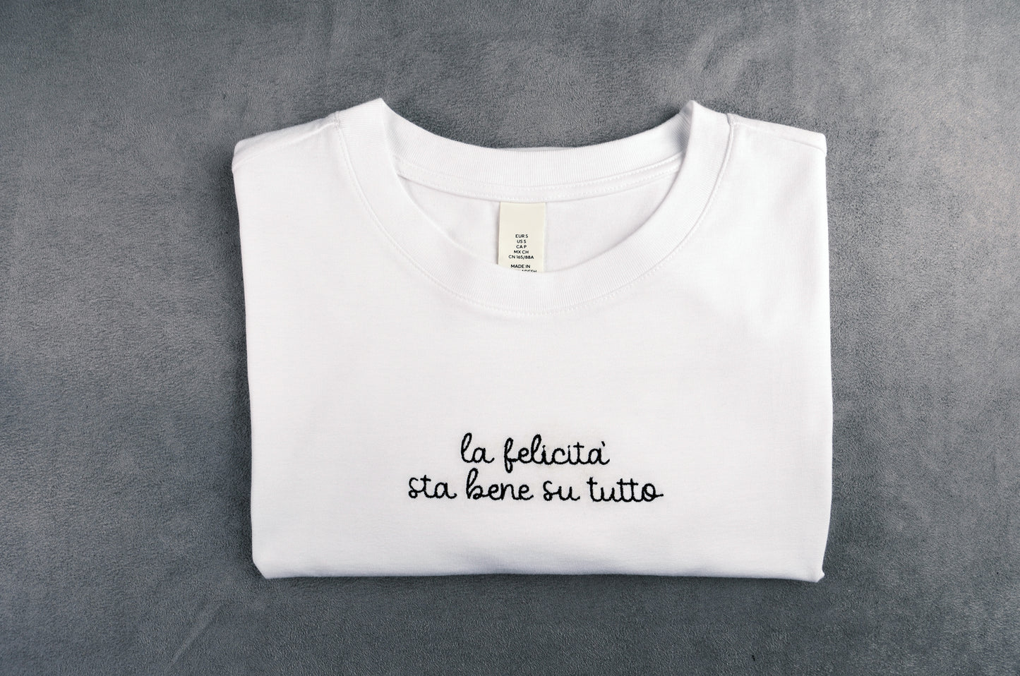 Dettaglio T-shirt “LA FELICITÀ STA BENE SU TUTTO”, la maglietta in 100% cotone è ricamata con la scritta "la felicità sta bene su tutto"